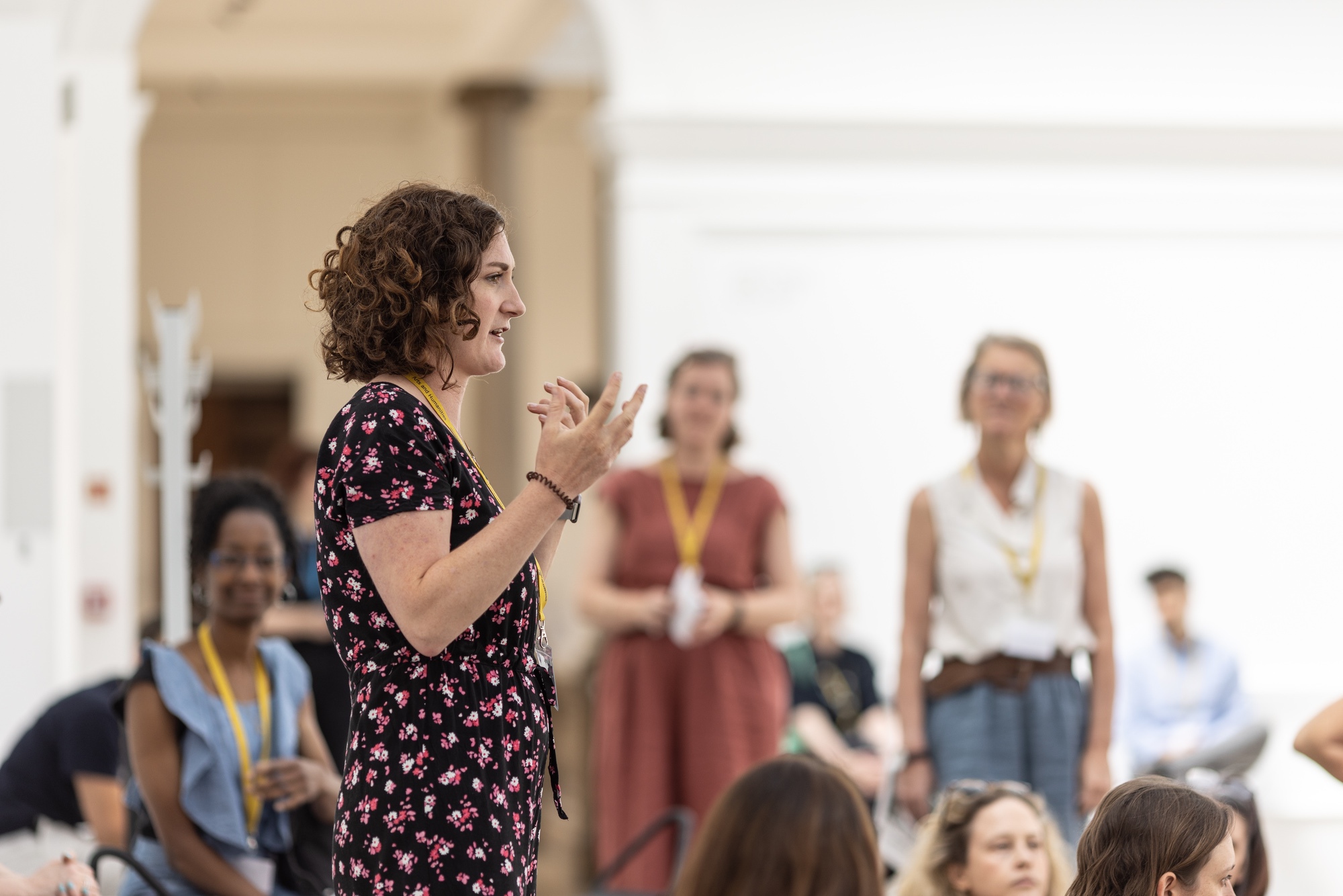 A person in the foreground side-on speaking to a group of people sitting and standing in the background